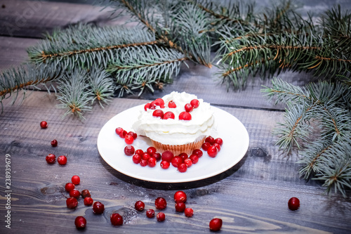 Freshly baked cake on a white plate decorated with red berries and whipped white cream with a plate topped with red berries decorated with spruce sprigs on a light wooden table top.