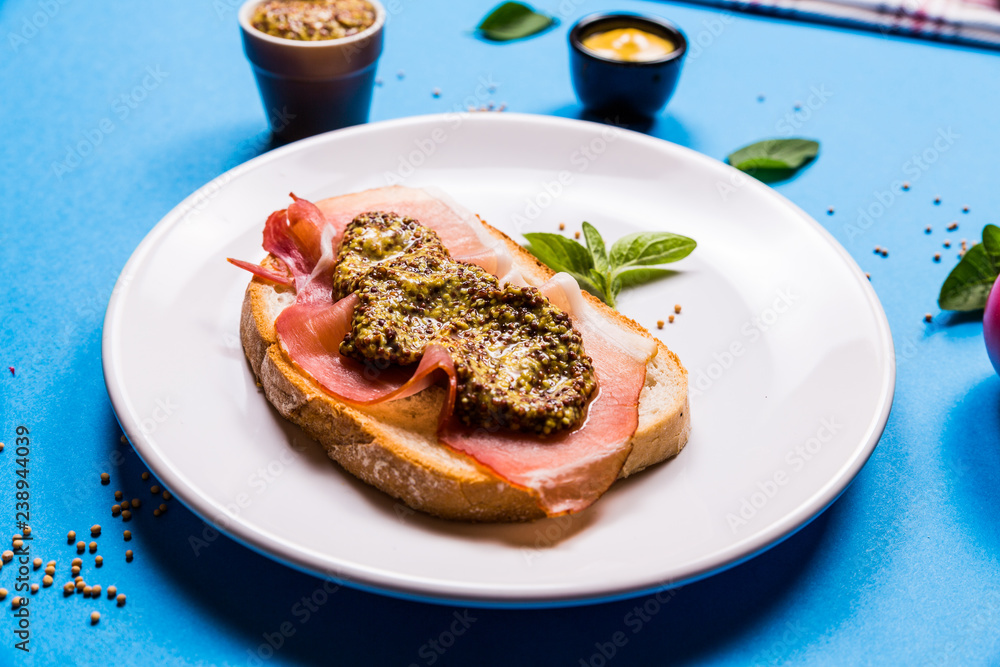 Sandwich with ham and mustard on a white ceramic plate, blue background Sandwich with ham and mustard on a white ceramic plate, blue background