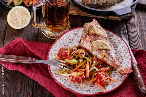 Delicious and healthy baked ocean perch in a plate with slices of lemon and rosemary next to grilled vegetables and light beer mug are ready to eat
