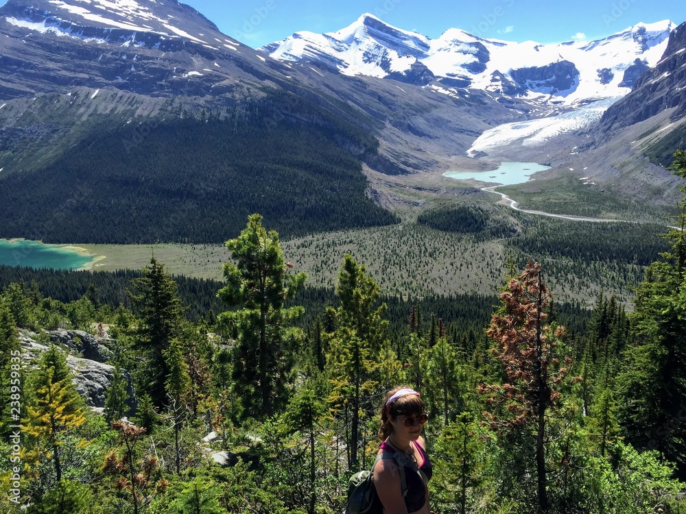Naklejka premium A young female hiker resting along the Berg Lake trail in British Columbia, Canada. Mount Robson and Berg Lake are surrounded by forests and mountains in the background