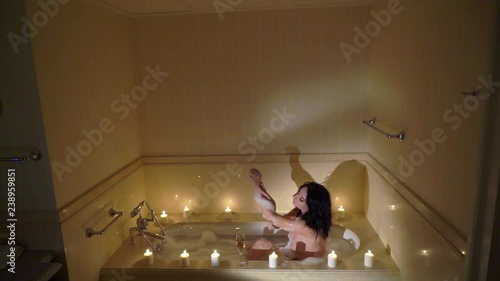 Young sexy brunette woman going to bathroom, undressing removing bathrobe and laying in bathtube with candles. Romantic dating. Slowmotion. Girl drinking champagne form a glass. photo