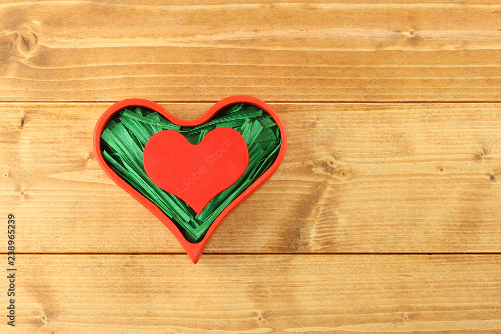 Red painted wooden heart enclosed with green paper raffia strips in red box isolated on wooden background Red painted wooden heart enclosed with green paper raffia strips in red box isolated on wooden background