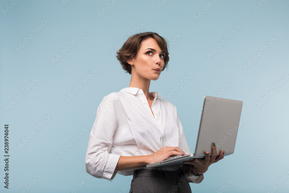 Young pretty businesswoman with dark short hair in white shirt holding laptop in hands while dreamily looking aside over blue background Young pretty businesswoman with dark short hair in white shirt holding laptop in hands while dreamily looking aside over blue background