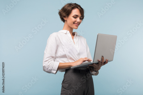 Young beautiful businesswoman with dark short hair in white shirt happily working on laptop over blue background isolated