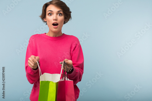 Young shocked woman with dark short hair in pink sweater holding colorful shopping bags in hands while amazedly looking in camera over blue background photo