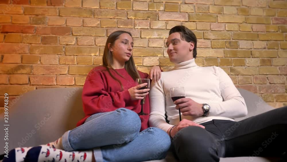 Portrait of young caucasian couple sitting on sofa and drinking wine communicating with each other in cosy home atmosphere. Portrait of young caucasian couple sitting on sofa and drinking wine communicating with each other in cosy home atmosphere.