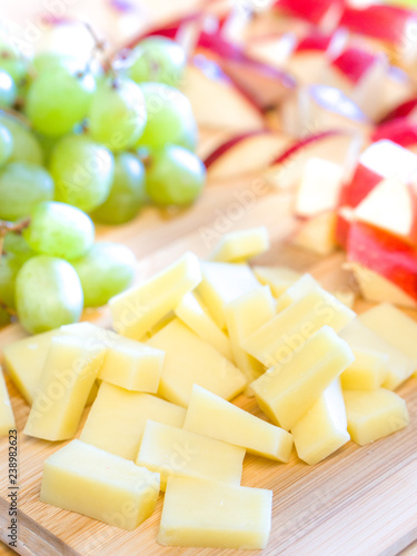 Close up photograph of a bamboo wood cutting board with an appetizer spread including cheese, green grapes and pieces of apple sliced up.