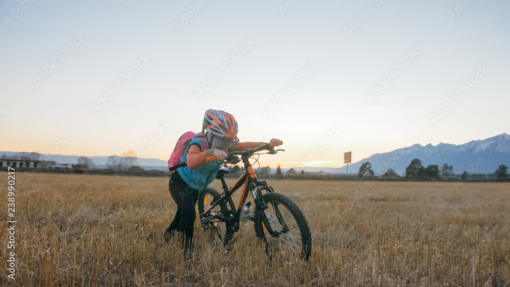 Obraz premium One caucasian children walk with bike in wheat field. Little girl walking black orange cycle on background of beautiful snowy mountains. Biker stand with backpack and helmet. Mountain bike hardtail.