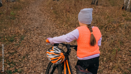 One caucasian children walk with bike in autumn park. Little girl walking black orange cycle in forest. Kid goes do bicycle sports. Biker motion ride with backpack and helmet. Mountain bike hardtail.