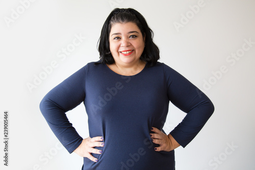 Smiling overweight Hispanic woman posing with hands on hips. Successful adult woman standing with akimbo gesture. Isolated on white. Success concept