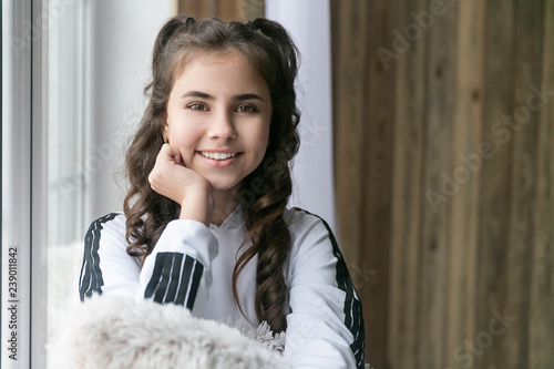 beautiful brunette girl sitting on the windowsill hugging a soft beige pillow and looking at the camera and smiling