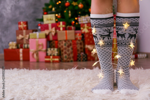 Cropped shot of female legs standing with lights wrapped around, wearing mid-calf socks with reindeer print and ornament, Decorated christmas tree on background. Copy space, close up.
