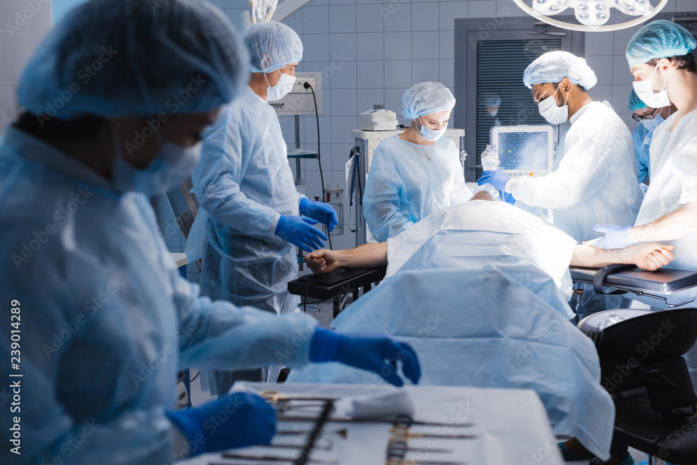 Asian female nurse checking all surgical instruments on tray on foreground. Mature male surgeon treating patient during surgical operation on background. Asian female nurse checking all surgical instruments on tray on foreground. Mature male surgeon treating patient during surgical operation on background.