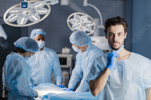 Portrait of handsome caucasian bearded young surgeon looking at camera with colleagues on background while standing in surgery with folded arms.