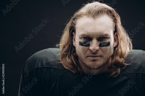 Face and shoulder portrait of confident American football a prolific goal scorer with painted black stripes and long blonde hair, looking at camera against black background