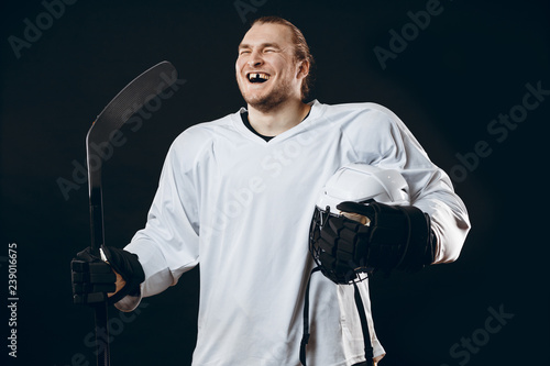 Happy Handsome hockey player with one broken front tooth laughing at camera, standing with stick in white uniform, isolated on black