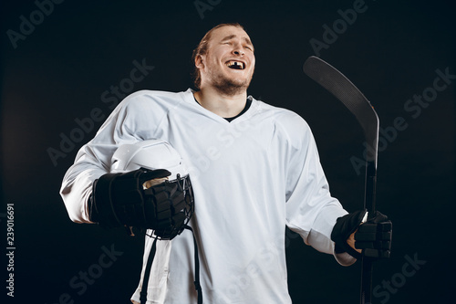 Happy Handsome hockey player with one broken front tooth laughing at camera, standing with stick in white uniform, isolated on black