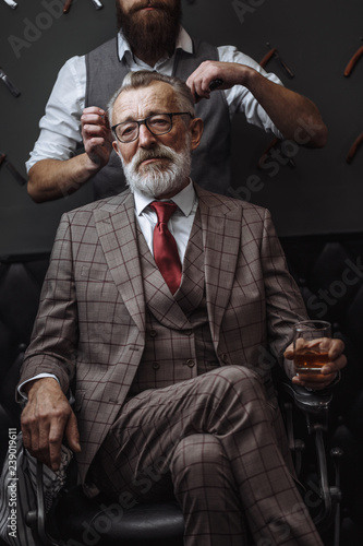 Professional male barber cutting hair of grey- haired wealthy man in years, dressed in fashionable three-piece suite. Process of creation of new hairdo in a luxury barbershop. © alfa27