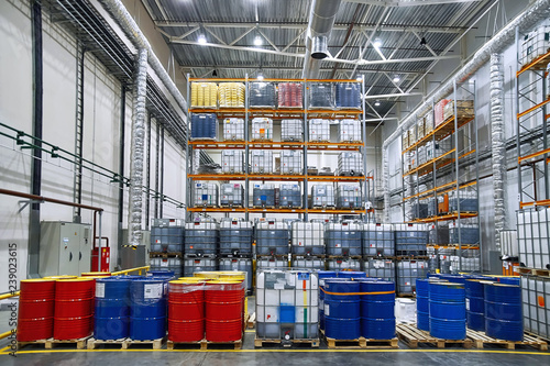 Oil drums and plastic container on pallets in a warehouse on metal shelving. Handling and storing industrial lubricants. Hazardous material storage. Red and blue tank