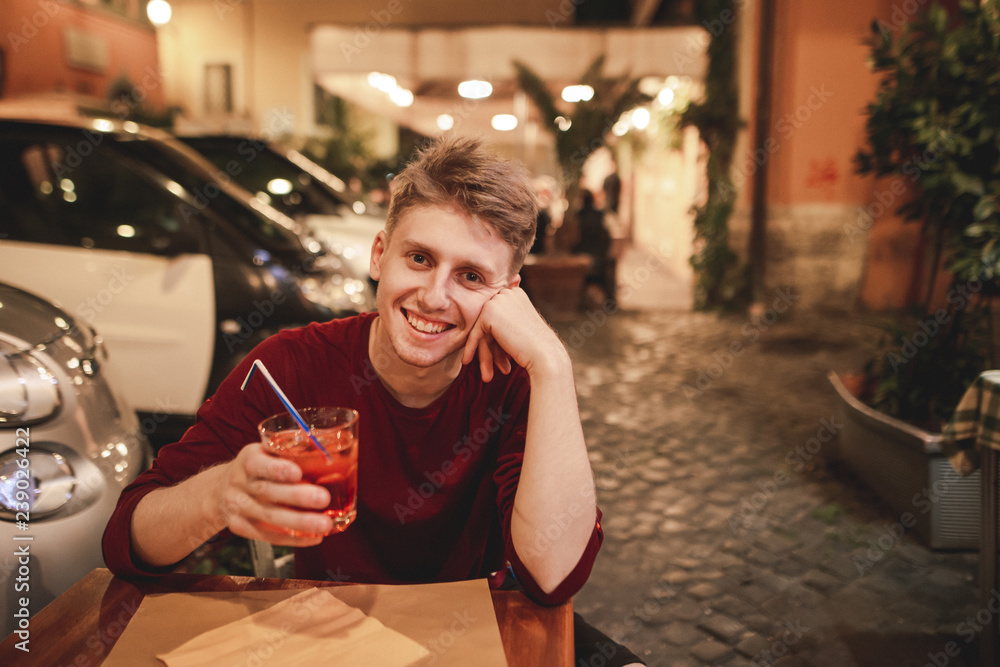 Φωτογραφία Portrait of a smiling young man who sits in the evening in a restaurant with a glass of alcohol in his hands,looks camera