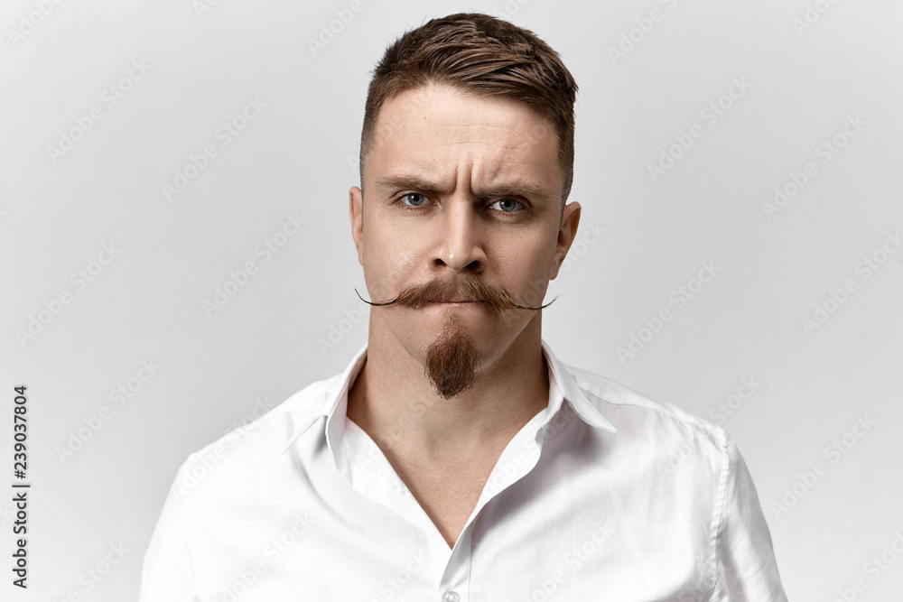 Close up shot of frustrated young man with stylish haircut, mustache and stubble frowning his eyebrows and pursing lips, having uneasy perplexed expression, being worried about problems at work Close up shot of frustrated young man with stylish haircut, mustache and stubble frowning his eyebrows and pursing lips, having uneasy perplexed expression, being worried about problems at work