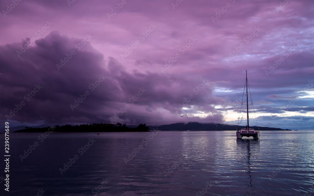 Lonely catamaran at sunset against the background of storm clouds near small islands in the Pacific ocean, French Polynesia.