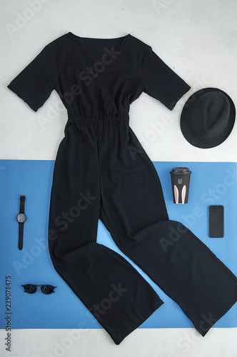 Unusual layout with stylish fashionable modern clothes. White and blue paper background. Black jumpsuit with various accessories and a cardboard cup for coffee.