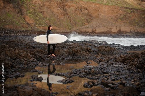 Guy standing with a surf in his hands on the rock beach