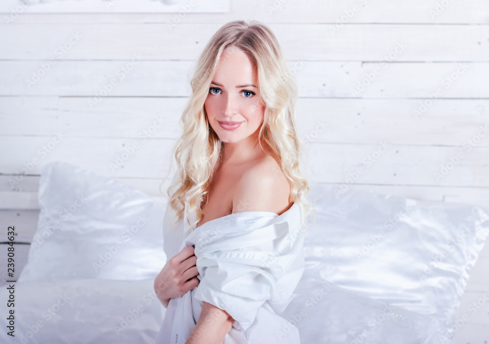 Young sexy blonde woman in a white shirt is sitting on a white bed in the studio
