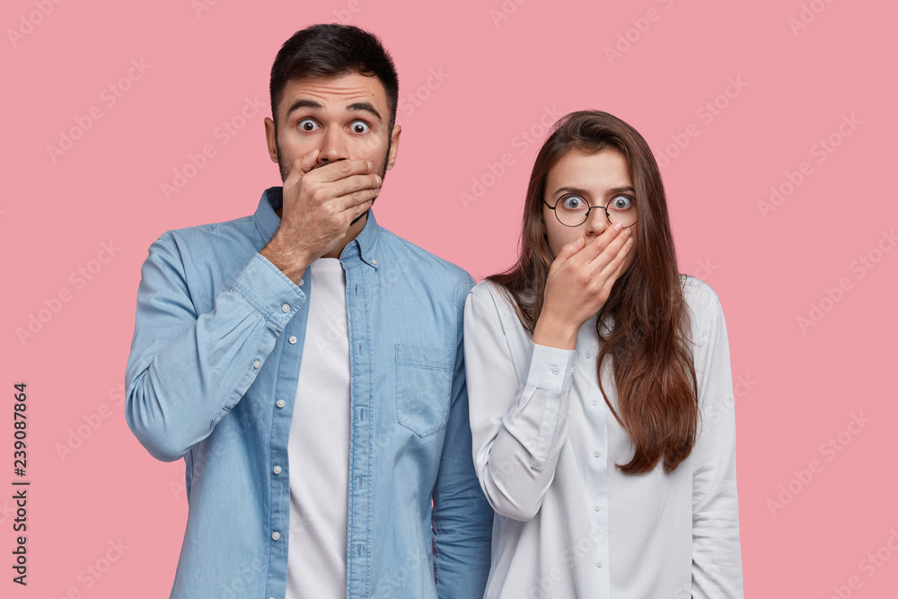Photo of scared speechless lady and man cover mouthes with palms, have stupefied expressions, dressed in elegant shirt, stand next to each other over pink studio wall. People, reaction and feeling Photo of scared speechless lady and man cover mouthes with palms, have stupefied expressions, dressed in elegant shirt, stand next to each other over pink studio wall. People, reaction and feeling
