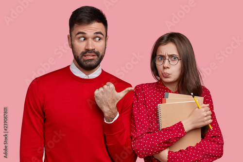 Handsome unshaven man wears red jumper, points at displeased beautiful woman who makes grimace, carries orgnaizer and spiral notepad, makes notes with pencil, suggests to ask her this question photo