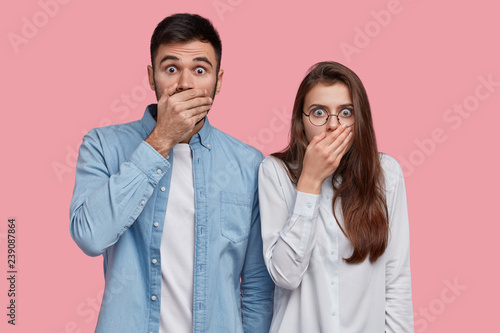 Photo of scared speechless lady and man cover mouthes with palms, have stupefied expressions, dressed in elegant shirt, stand next to each other over pink studio wall. People, reaction and feeling
