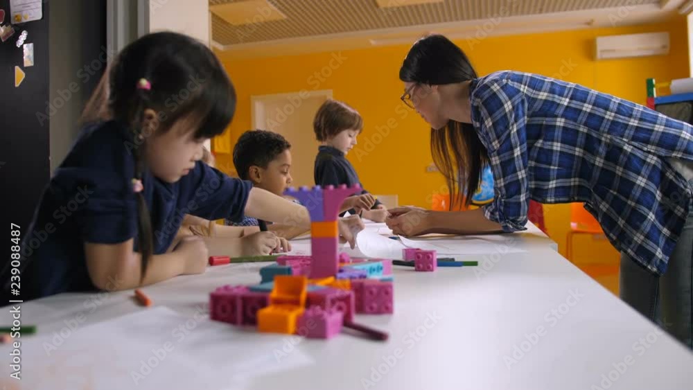 Beautiful female mixed race teacher and little mulitcultural students working together in craft lesson. Positive kindergarten teacher helping diverse kids with drawing and painting in preschool class. Beautiful female mixed race teacher and little mulitcultural students working together in craft lesson. Positive kindergarten teacher helping diverse kids with drawing and painting in preschool class.