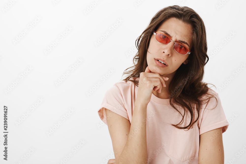 Portrait of confused unsure attractive young female in red sunglasses tilting head and holding hand on chin as looking focused and questioned at camera as observing something strange and confusing