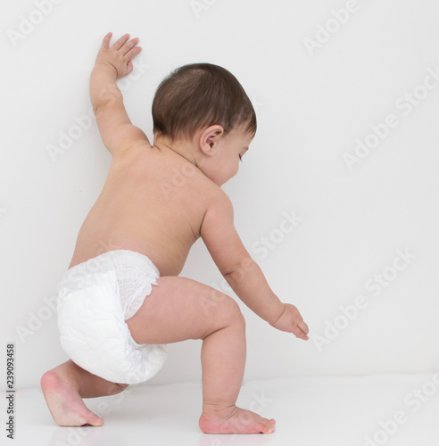 Baby wear white diaper try to stand up by push his left hand on the wall,  in white background