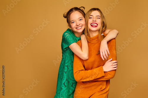 Two young beautiful blond smiling hipster girls posing in trendy summer clothes. Carefree women isolated on golden background. Positive models going crazy