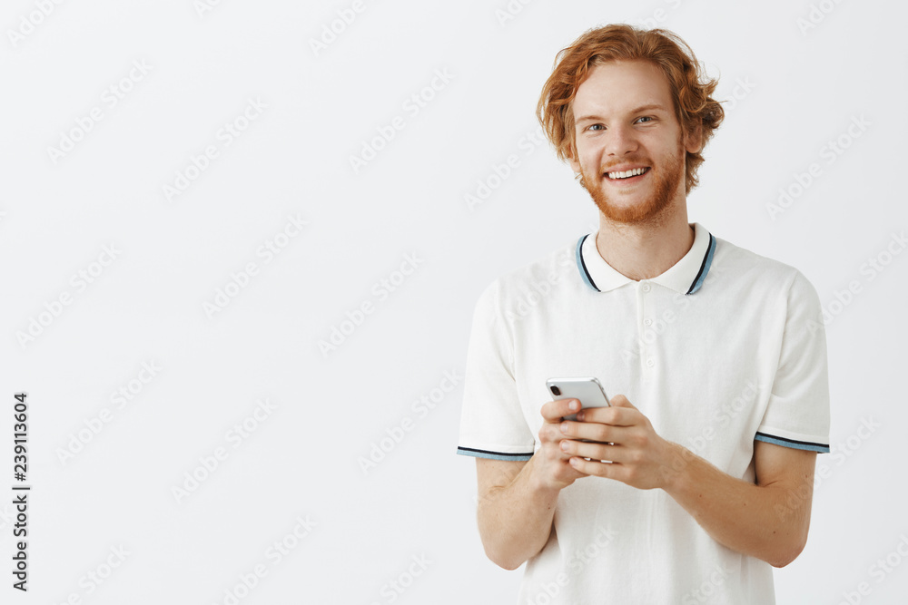 Waist-up shot of charming carefree ordinary urban redhead guy in casual outfit with beard holding smartphone in both hands and smiling joyfully trying new phone and checking workability of device Waist-up shot of charming carefree ordinary urban redhead guy in casual outfit with beard holding smartphone in both hands and smiling joyfully trying new phone and checking workability of device