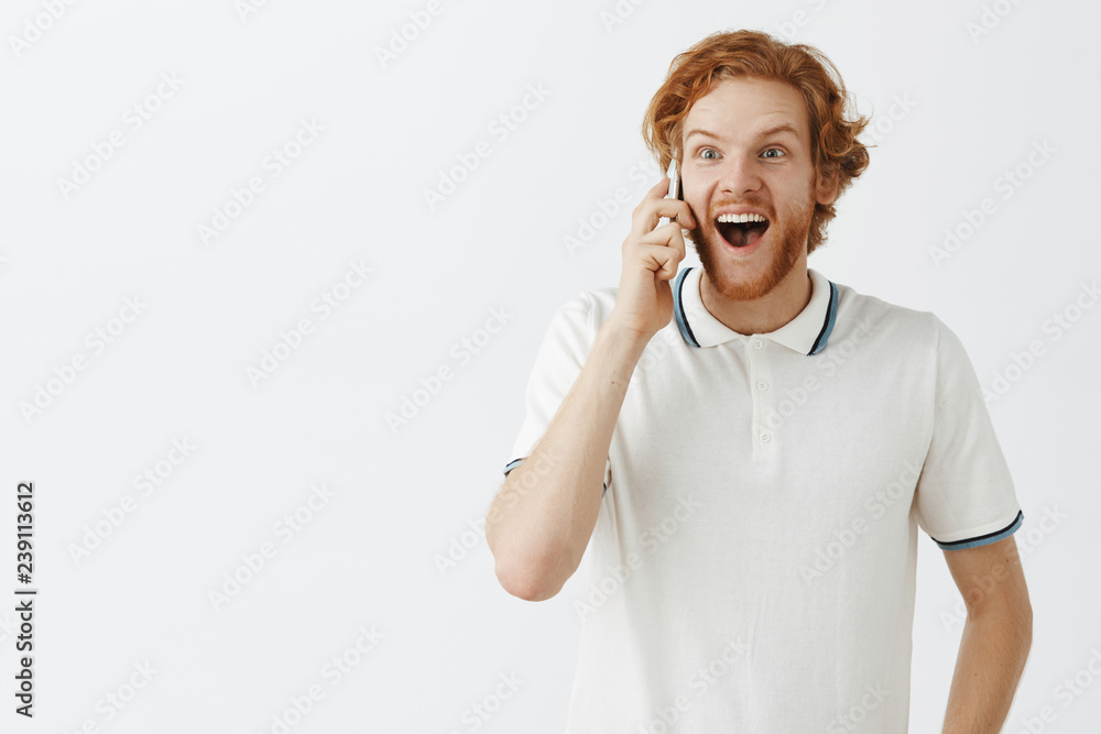 Waist-up shot of excited and overemotive good-looking young caucasian redhead male student in polo shirt with beard holding smartphone near ear and smiling broadly with amazement and thrill Waist-up shot of excited and overemotive good-looking young caucasian redhead male student in polo shirt with beard holding smartphone near ear and smiling broadly with amazement and thrill