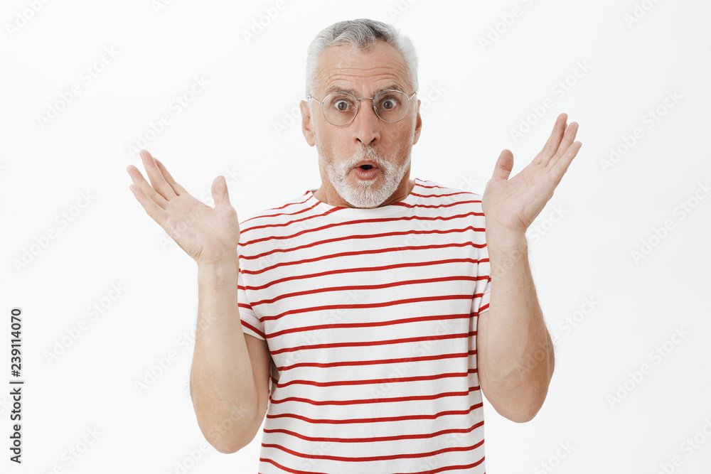 Waist-up shot of surprised handsome senior man with grey beard in prescribed glasses and striped t-shirt raising palms in amazement reacting on unxpected news opening mouth popping eyes at camera Waist-up shot of surprised handsome senior man with grey beard in prescribed glasses and striped t-shirt raising palms in amazement reacting on unxpected news opening mouth popping eyes at camera