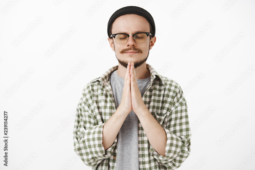 Pleased peaceful and calm handsome young male student in black beanie and checked shirt closing eyes and smiling with delight holding hands in pray talking to god or making wish over white background Pleased peaceful and calm handsome young male student in black beanie and checked shirt closing eyes and smiling with delight holding hands in pray talking to god or making wish over white background