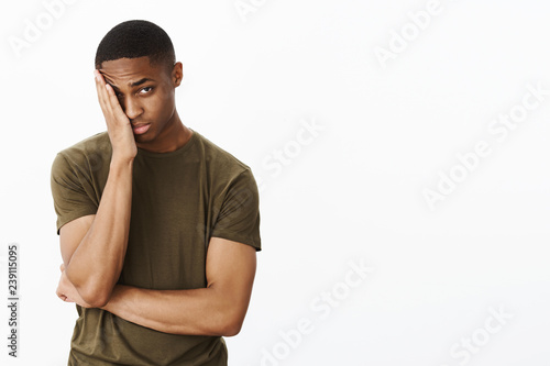 Portrait of man feeling embarrassed making facepalm gesture putting hand to half of face peeking at camera with tired blushing expression standing displeased and exhausted over gray background