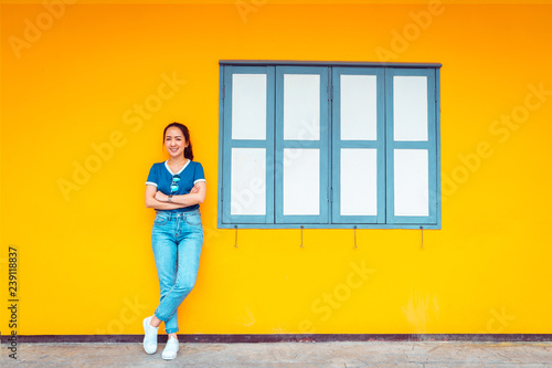 hipster girl standing beside yellow well and vintage window with posing relaxing smile by wearing blue shirt and blue jeans