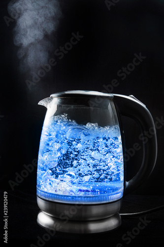 Boiling glass kettle with blue light on a black background, steam comes from the spout