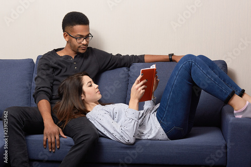 Photo of relaxed Caucasian young woman lies on knees of her dark skinned boyfriend, reads book aloud, pose together on comfortable sofa in living room, enjoy domestic atmopshere and coziness. photo