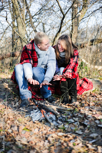 Young loving couple dressed in blue sweater sitting with red coffee cups on the firewood in winter forest