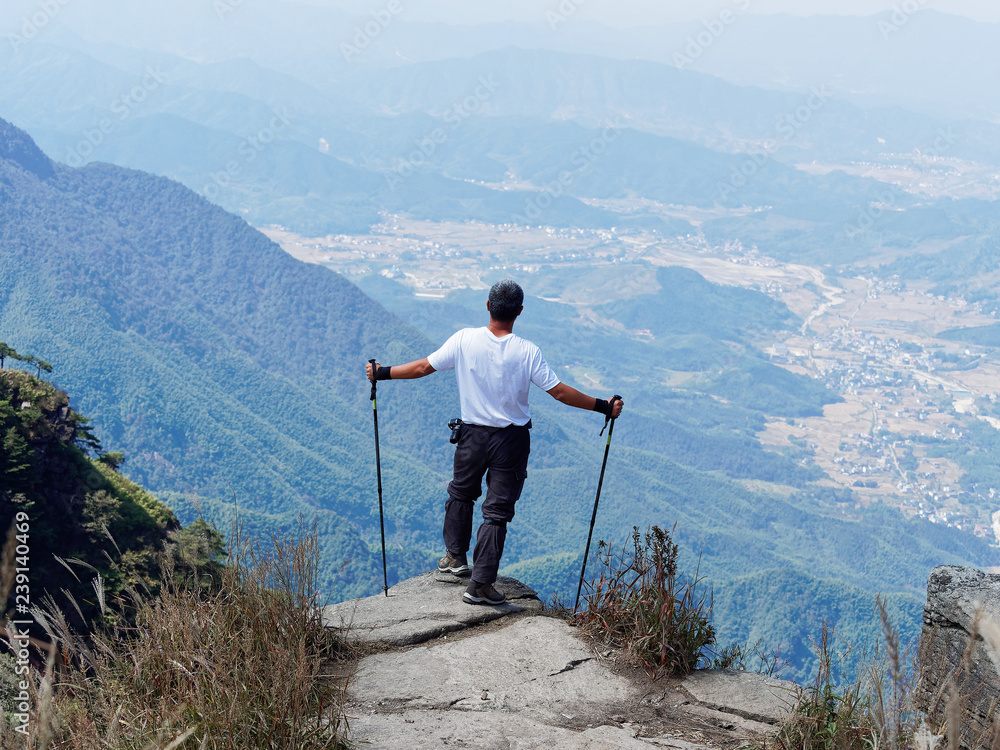 Mountain hiker looking into the landscape of town at the foot of the mountain. Rear view of the man stand on the top of the mountain cliff holding with two alpenstocks. 