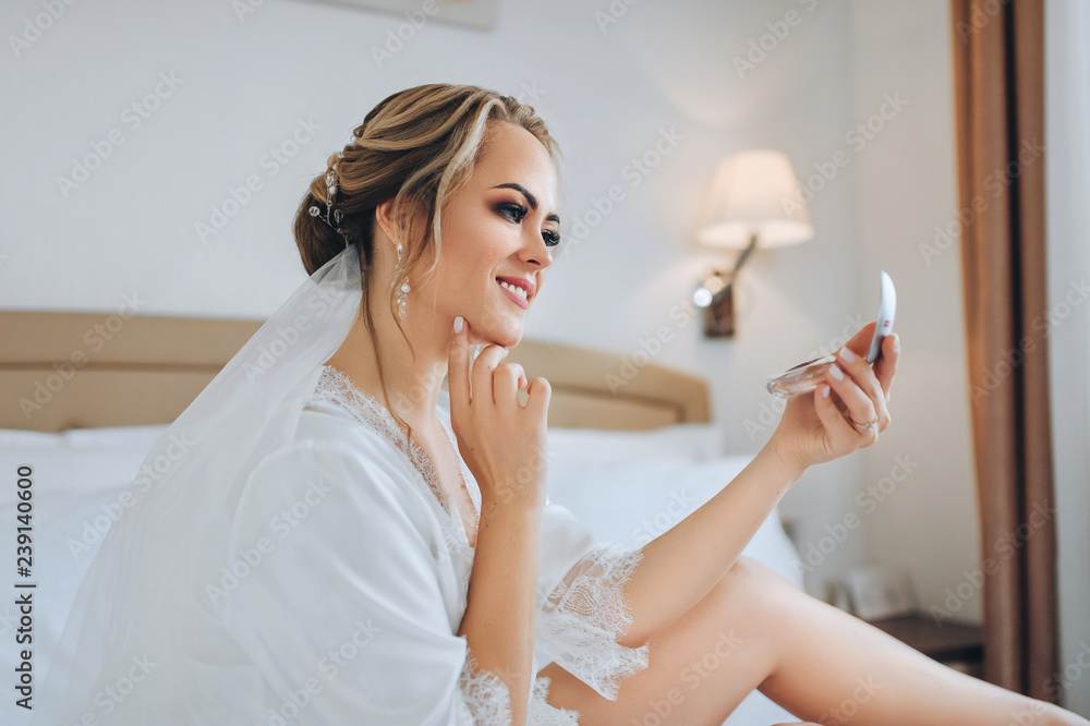 Beautiful bride sits on a white bed and looks in a pocket mirror. Wedding interior. Charges the bride at the hotel. Wedding photography. Portrait of a cute bride. Beautiful bride sits on a white bed and looks in a pocket mirror. Wedding interior. Charges the bride at the hotel. Wedding photography. Portrait of a cute bride.