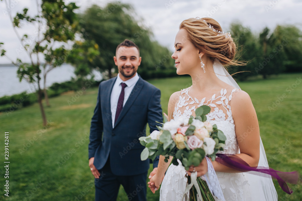 A blond and smiling bride with a bouquet and a bearded groom hold hands against the background of a park with green grass and willows. Wedding portrait of beautiful newlyweds. Bride profile. Wedding. A blond and smiling bride with a bouquet and a bearded groom hold hands against the background of a park with green grass and willows. Wedding portrait of beautiful newlyweds. Bride profile. Wedding.
