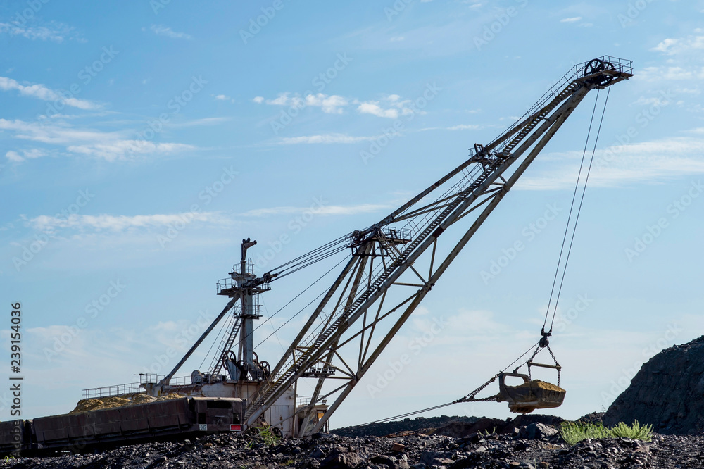 A huge excavator overloads in the dumps rock from the unloaded train from the mine against a clear blue sky. Concept: mining and environmental problems A huge excavator overloads in the dumps rock from the unloaded train from the mine against a clear blue sky. Concept: mining and environmental problems