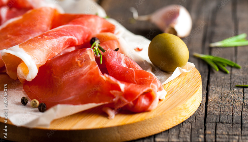 Closeup of thin slices of prosciutto with mixed olives and paprika on wooden cutting board Closeup of thin slices of prosciutto with mixed olives and paprika on wooden cutting board